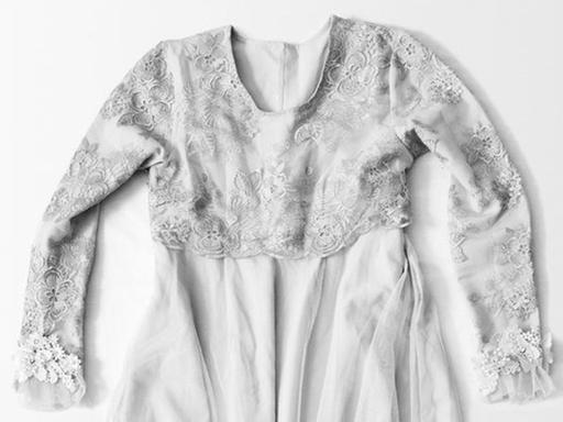 Ein weißes, bodenlanges Kleid, bestehend aus einem schlichten Rock und einem langärmeligen Oberteil aus Blumenspitze