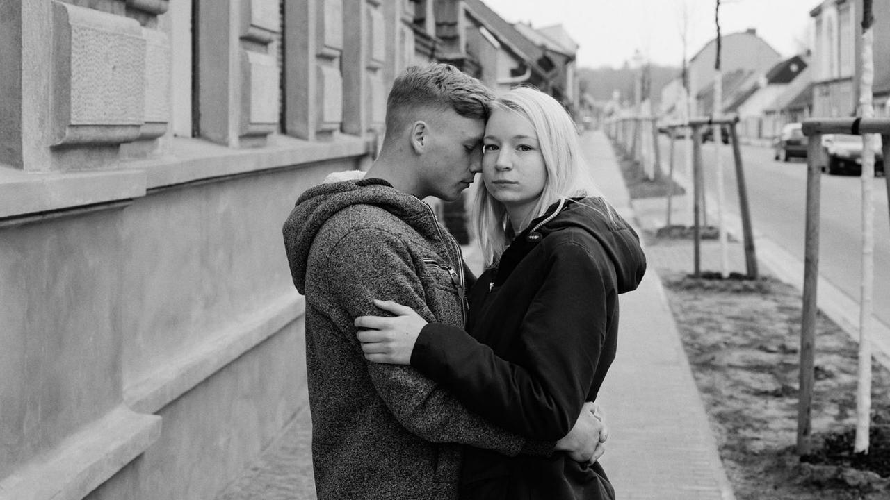 Ein schwarz-weiß Fotografie zeigt ein junges Paar mit den Köpfen aneinander gelehnt in einer Straßenszene in einer ostdeutschen Kleinstadt.