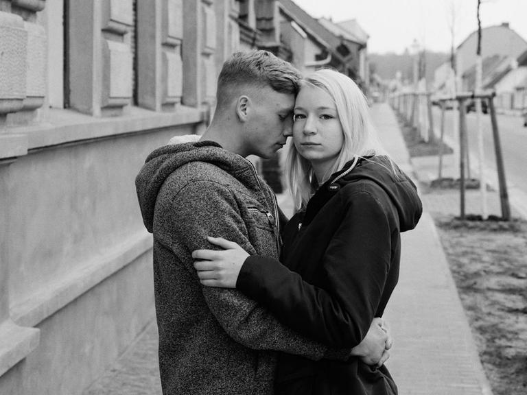 Ein schwarz-weiß Fotografie zeigt ein junges Paar mit den Köpfen aneinander gelehnt in einer Straßenszene in einer ostdeutschen Kleinstadt.