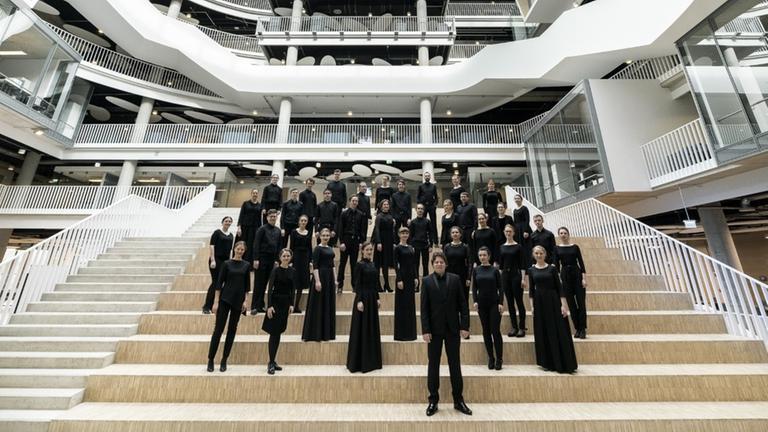 Der Chor und sein Leiter stehen in schwarzer Konzertkleidung in einem modernen, weißen Raum.