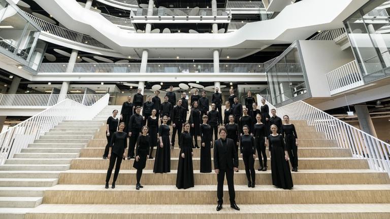 Der Chor und sein Leiter stehen in schwarzer Konzertkleidung in einem modernen, weißen Raum. Der Chor und sein Leiter stehen in schwarzer Konzertkleidung in einem modernen, weißen Raum.