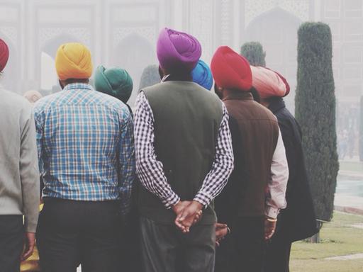 Zu sehen sind sechs Männer in der Rückenansicht in einem Park, die alle einen Turban in unterschiedlichen bunten Farben tragen