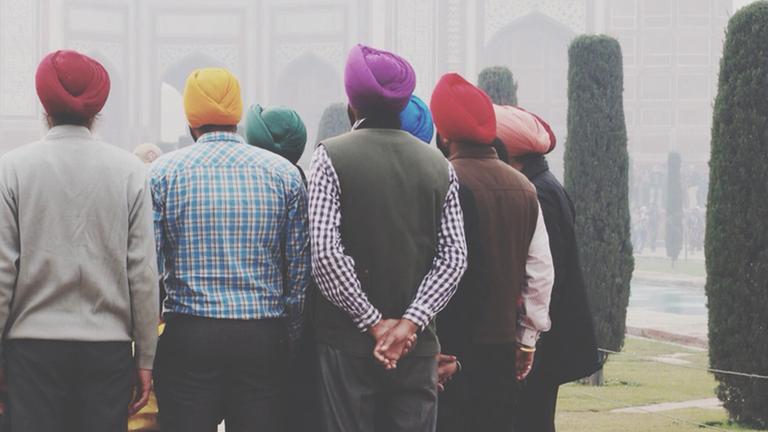 Zu sehen sind sechs Männer in der Rückenansicht in einem Park, die alle einen Turban in unterschiedlichen bunten Farben tragen