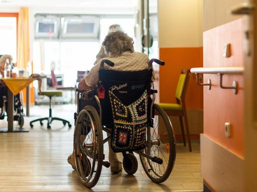 Eine Frau im Rollstuhl sitzt allein im Gemeinschaftsraum eines Altenheims.