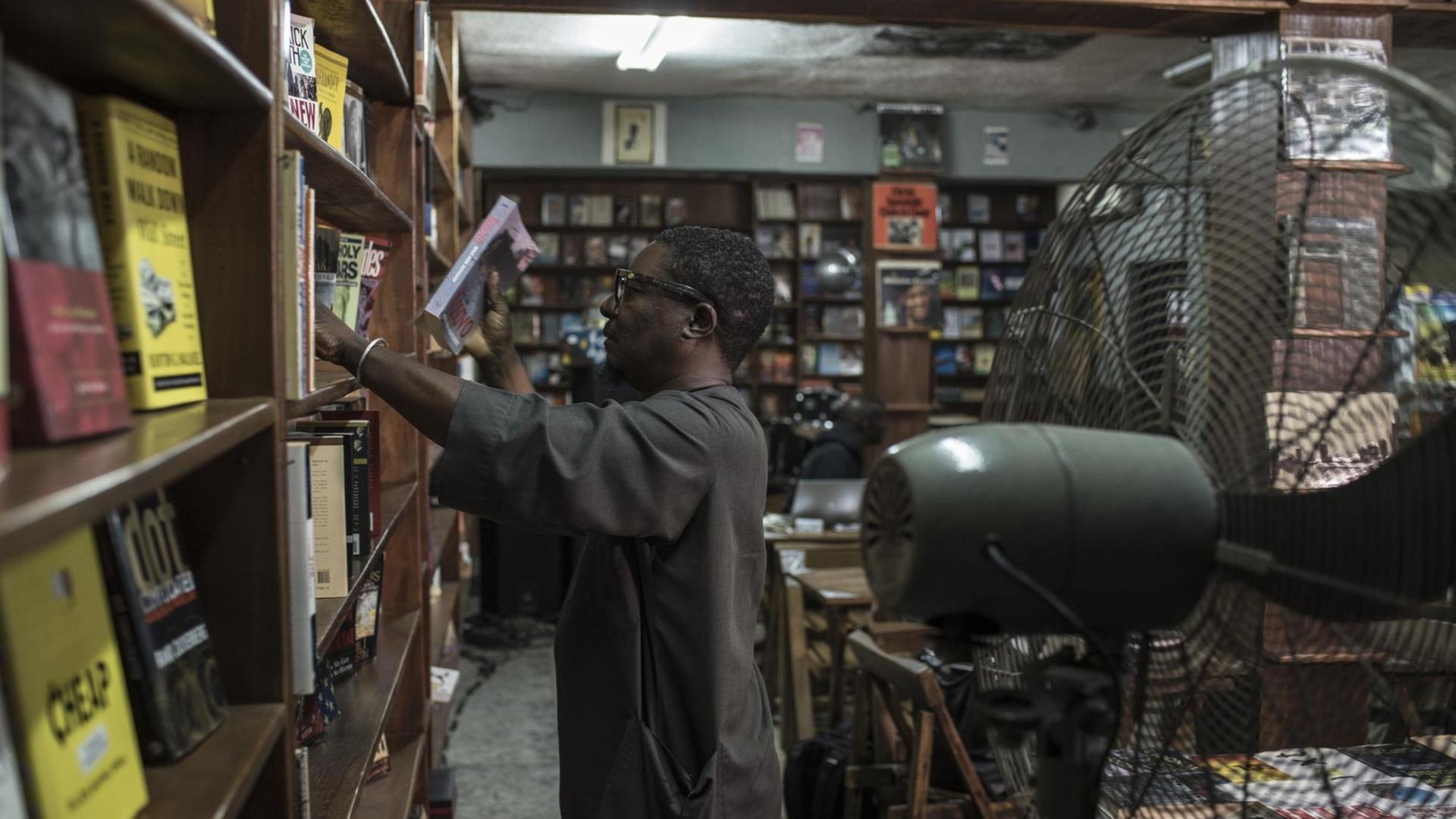 Der Inhaber von The Jazz Hole, Kunle Tejuoso, ordnet die deckenhohen Bücherregale in seinem Laden, während ein großer Ventilator für Abkühlung sorgt. The Jazz Hole ist ein unabhängiger Platten- und Buchladen in Lagos.