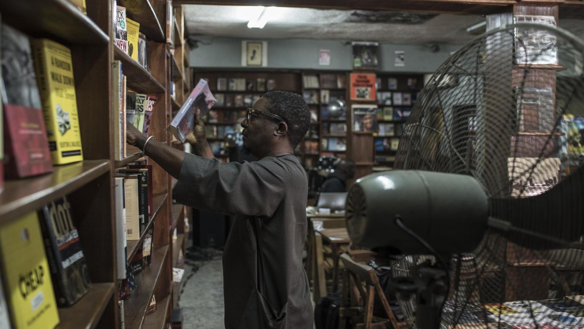 Der Inhaber von The Jazz Hole, Kunle Tejuoso, ordnet die deckenhohen Bücherregale in seinem Laden, während ein großer Ventilator für Abkühlung sorgt. The Jazz Hole ist ein unabhängiger Platten- und Buchladen in Lagos. Der Inhaber von The Jazz Hole, Kunle Tejuoso, ordnet die deckenhohen Bücherregale in seinem Laden, während ein großer Ventilator für Abkühlung sorgt. The Jazz Hole ist ein unabhängiger Platten- und Buchladen in Lagos.
