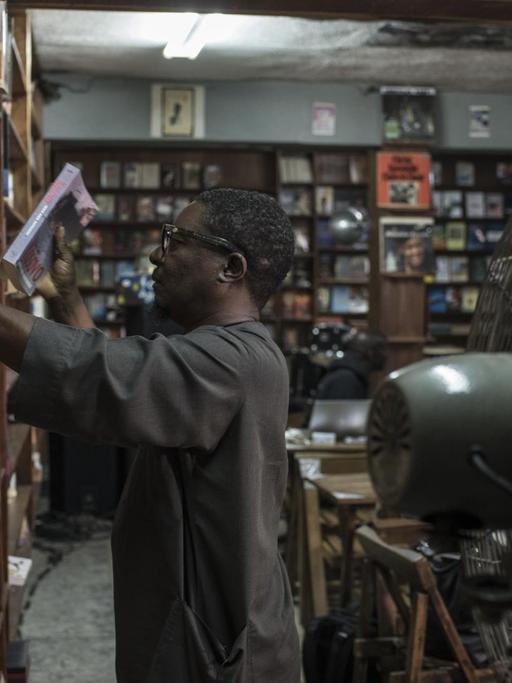 Der Inhaber von The Jazz Hole, Kunle Tejuoso, ordnet die deckenhohen Bücherregale in seinem Laden, während ein großer Ventilator für Abkühlung sorgt. The Jazz Hole ist ein unabhängiger Platten- und Buchladen in Lagos.