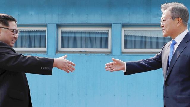 Die beiden schreiten vor einer blauen Hauswand lächelnd und mit vorgestreckten Händen aufeinander zu.
