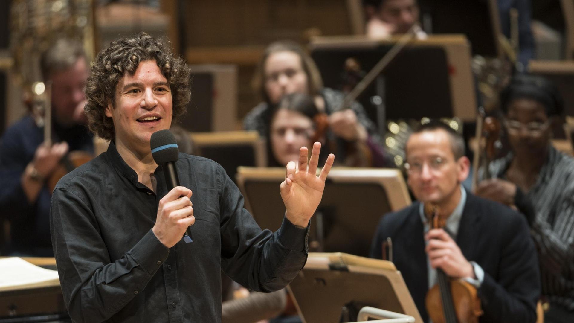 Robin Ticciati steht mit Mikrofon im Vordergrund, im Hintergrund sieht man das Deutsche Symphonie-Orchester Berlin.