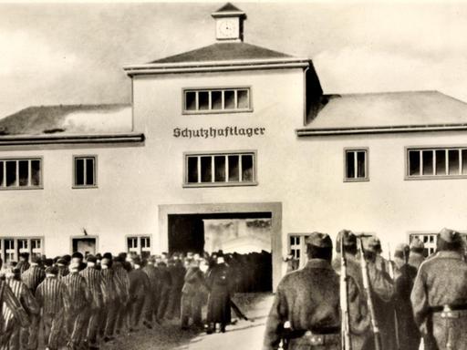Häftlinge und Wärter stehen vor dem Eingang zum KZ Sachsenhausen. Über dem Tor der Schriftzug: Schutzhaftlager.