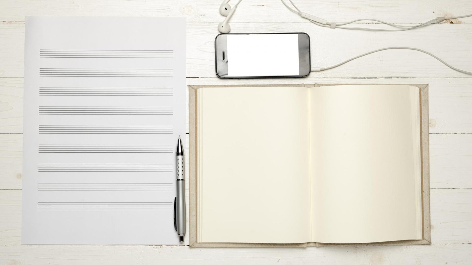 Leeres Notenpapier liegt auf einem hellen Holztisch. Daneben liegen ein Kugelschreiben, ein leeres Notizbuch und ein Smartphone mit Kopfhörern.