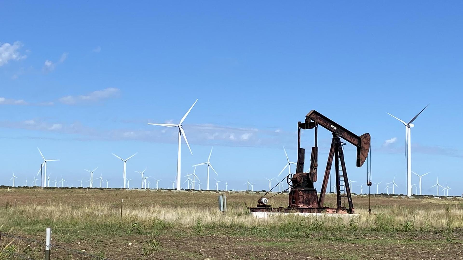 Auf einem flachen Acker steht im Vordergrund eine rostige Ölpumpe und im Hintergrund viele weiße Windräder. Auf einem flachen Acker steht im Vordergrund eine rostige Ölpumpe und im Hintergrund viele weiße Windräder.