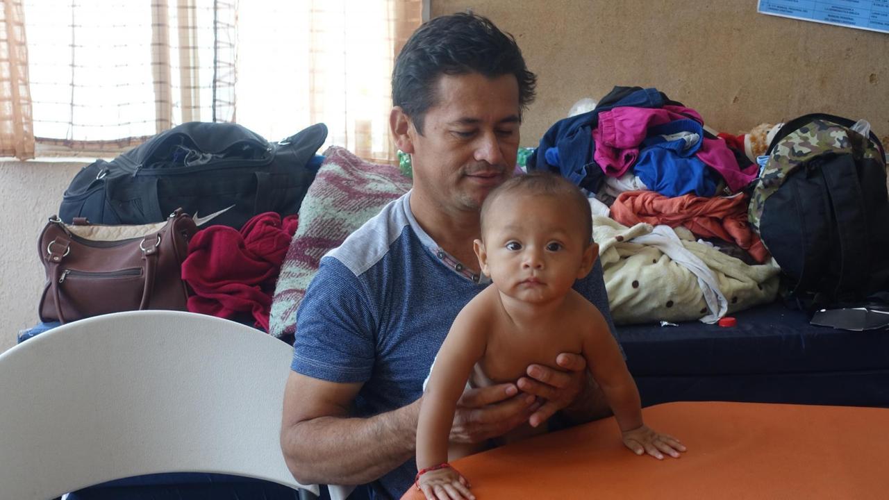 Mann mit Baby, im Hintergrund Klamottenberge auf einer Pritsche Mann mit Baby, im Hintergrund Klamottenberge auf einer Pritsche