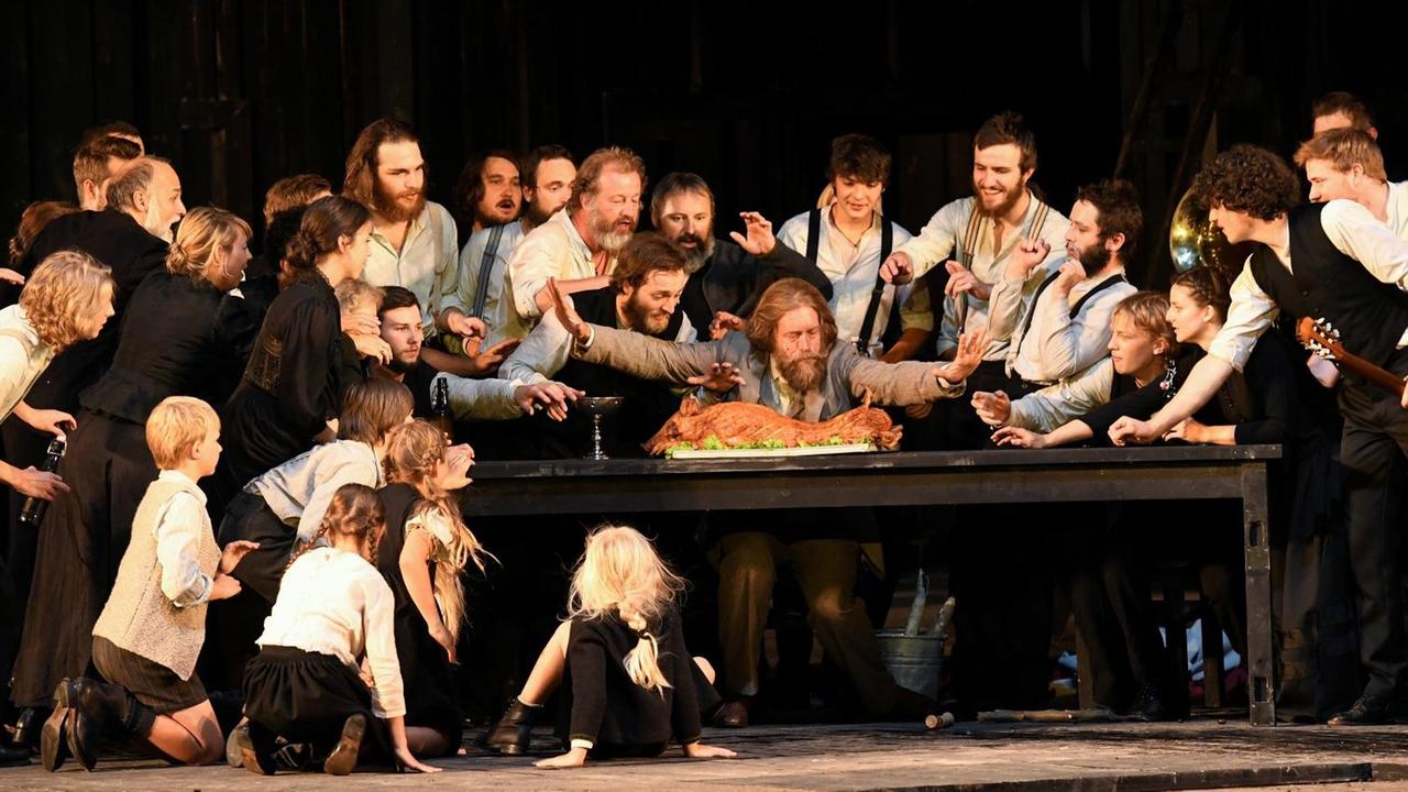 Ein Schauspielensemble stellt auf einer Theaterbühne eine Dorfgemeinschaft aus Männern, Frauen und Kindern dar. Alle drängen sich um einen Tisch auf dem ein ganzes zubereitetes Schwein liegt. Ein Schauspielensemble stellt auf einer Theaterbühne eine Dorfgemeinschaft aus Männern, Frauen und Kindern dar. Alle drängen sich um einen Tisch auf dem ein ganzes zubereitetes Schwein liegt.