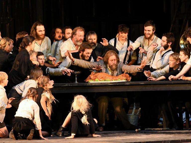 Ein Schauspielensemble stellt auf einer Theaterbühne eine Dorfgemeinschaft aus Männern, Frauen und Kindern dar. Alle drängen sich um einen Tisch auf dem ein ganzes zubereitetes Schwein liegt.