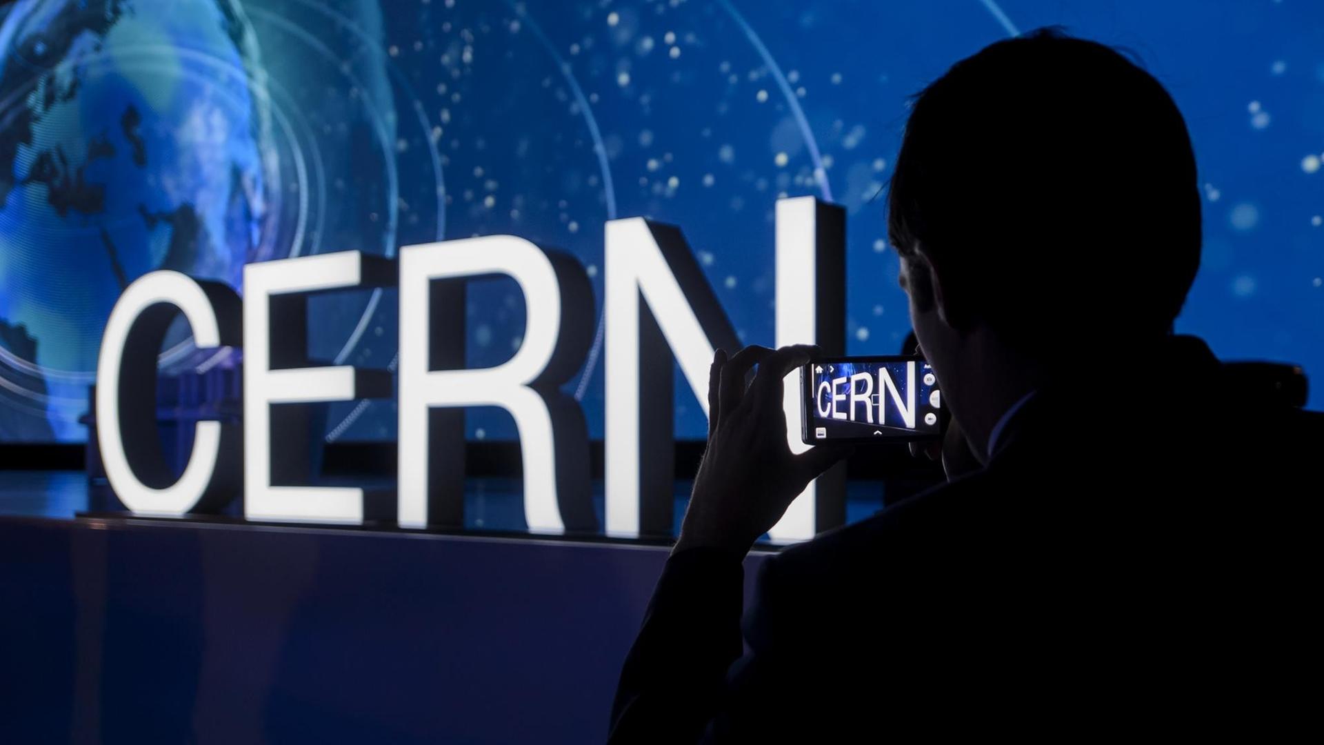 Beleuchteter Schriftzug "Cern" an einer dunklen Wand, rechts im Vordergrund der Schatten einer Person, die den Schriftzug fotografiert Beleuchteter Schriftzug "Cern" an einer dunklen Wand, rechts im Vordergrund der Schatten einer Person, die den Schriftzug fotografiert