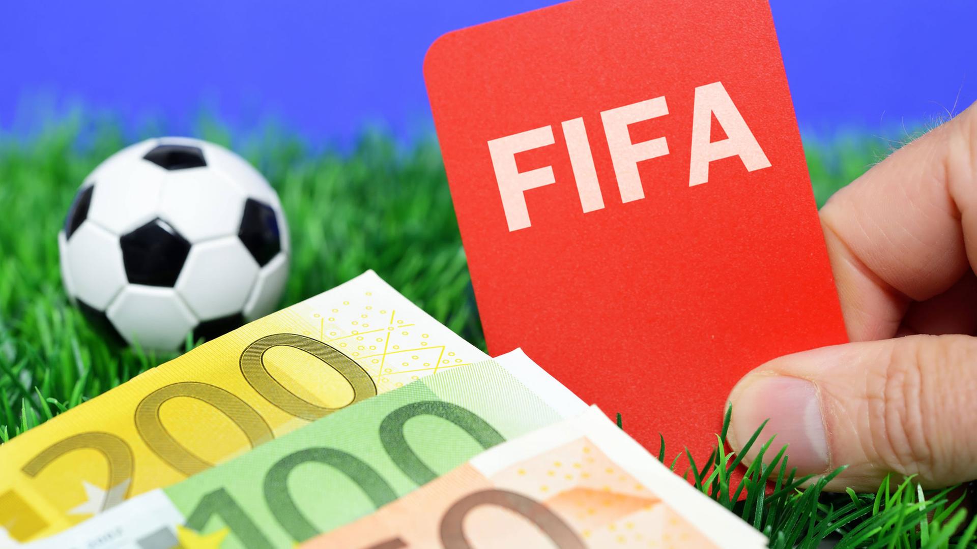 Ein Miniaturfußball auf einer Wiese neben einer Roten Karte mit FIFA-Schriftzug und mehreren Geldscheinen. Ein Miniaturfußball auf einer Wiese neben einer Roten Karte mit FIFA-Schriftzug und mehreren Geldscheinen.