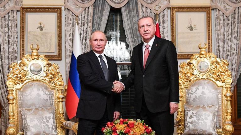 Putin und Erdogan stehen in einem pompös eingerichteten Raum hinter einem Tisch und zwischen zwei mit Gold verzierten Stühlen und schütteln sich die Hände. Putin und Erdogan stehen in einem pompös eingerichteten Raum hinter einem Tisch und zwischen zwei mit Gold verzierten Stühlen und schütteln sich die Hände.