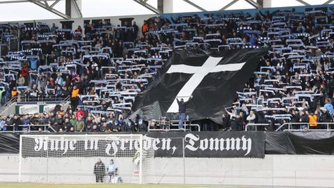 Im Fanblock des FC Chemnitz ist eine schwarze Fahne mit einem Kreuz aufgespannt. Darunter auf einem weiteren Banner "Ruhe in Frieden, Tommy"