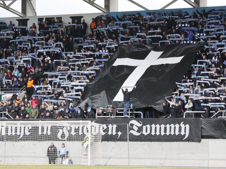 Im Fanblock des FC Chemnitz ist eine schwarze Fahne mit einem Kreuz aufgespannt. Darunter auf einem weiteren Banner "Ruhe in Frieden, Tommy"