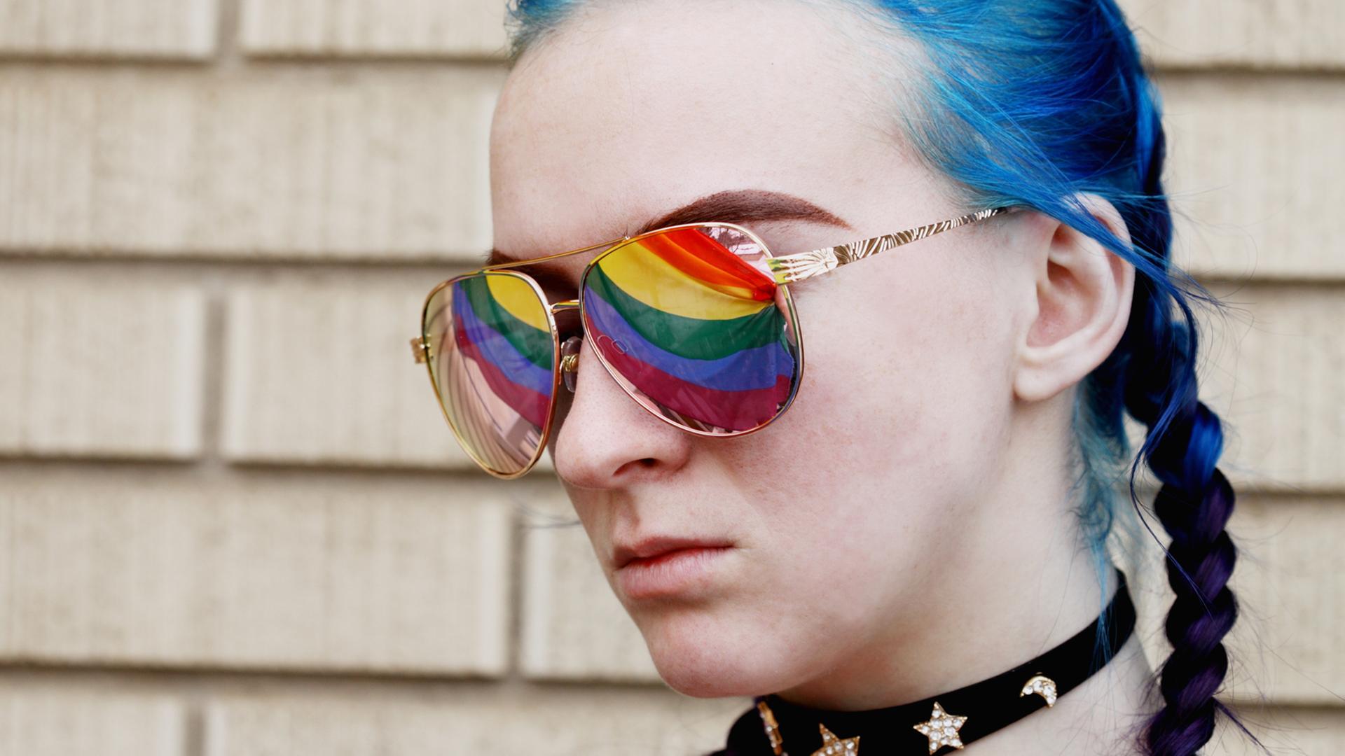 Eine junge Frau mit blau gefärbtem Haar trägt eine Sonnenbrille, in der sich die Regenbogenfahne spiegelt.