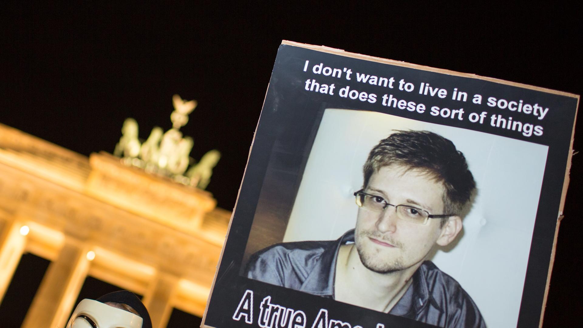Ein Demonstrant der Gruppe Anonymous steht vor dem Brandenburger Tor bei Nacht und hält ein Plakat mit einem Foto von Edward Snowden in die Höhe. Ein Demonstrant der Gruppe Anonymous steht vor dem Brandenburger Tor bei Nacht und hält ein Plakat mit einem Foto von Edward Snowden in die Höhe.