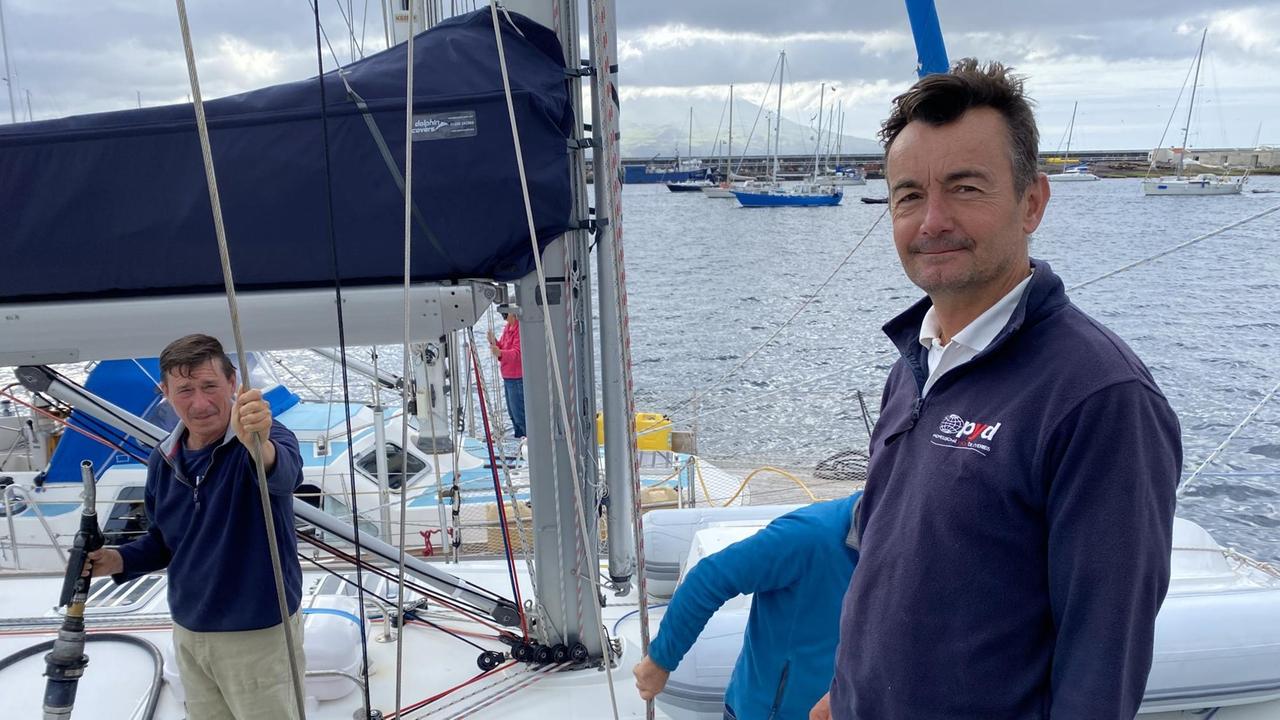 Ein älterer Mann mit blauem Pulli steht vor einer Segel-Yacht am Ufer, auf der eine Crew arbeitet. Ein älterer Mann mit blauem Pulli steht vor einer Segel-Yacht am Ufer, auf der eine Crew arbeitet.