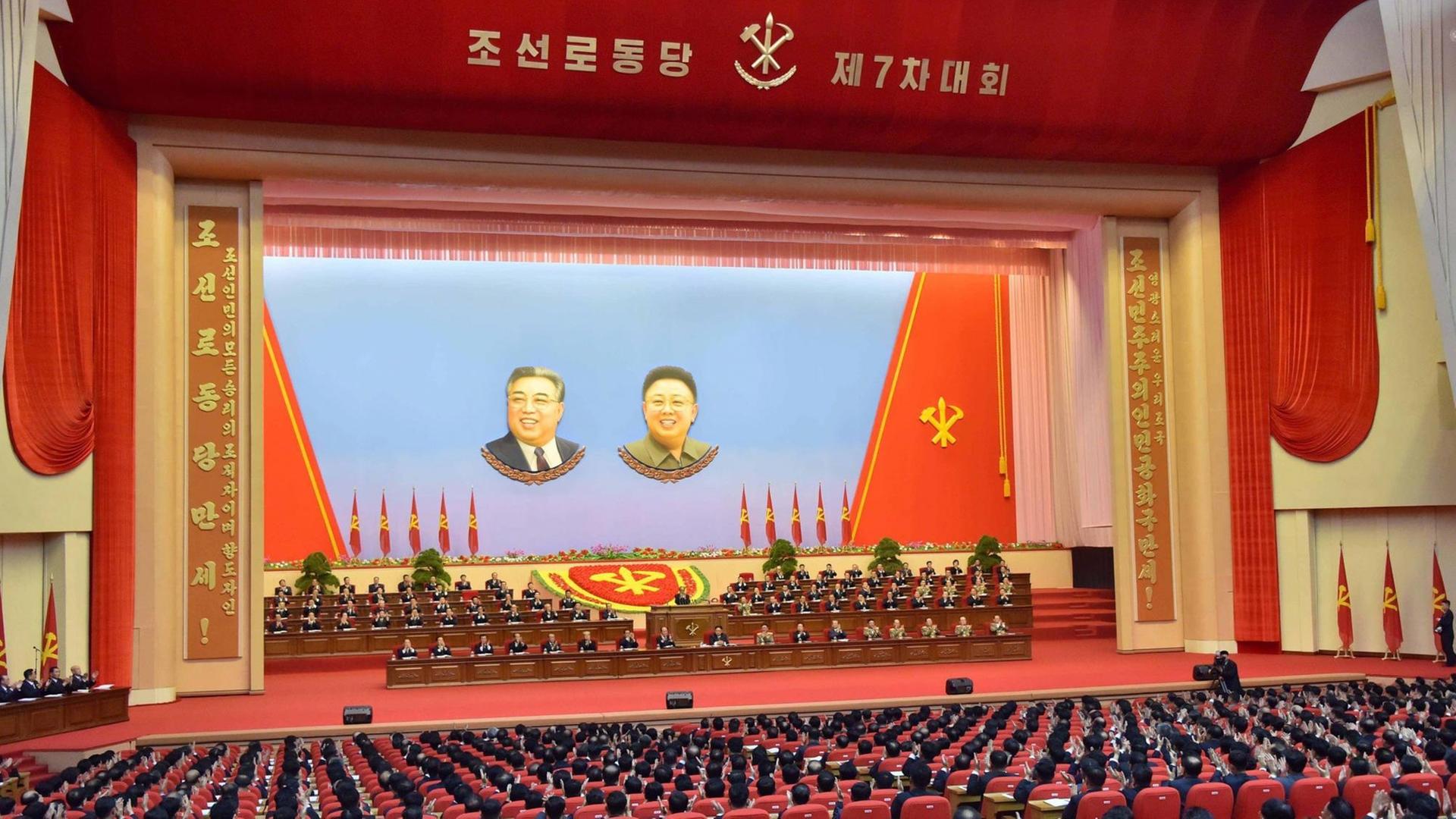 Ein großer Saal mit Bühne, die von einem roten Umhang gesäumt ist. In der Bühne hängen große Bilder von Kim Jong-un und Kim Il-sung, auf der Bühen sitzen Parteifunktionäre, im Publikum Delegierte.