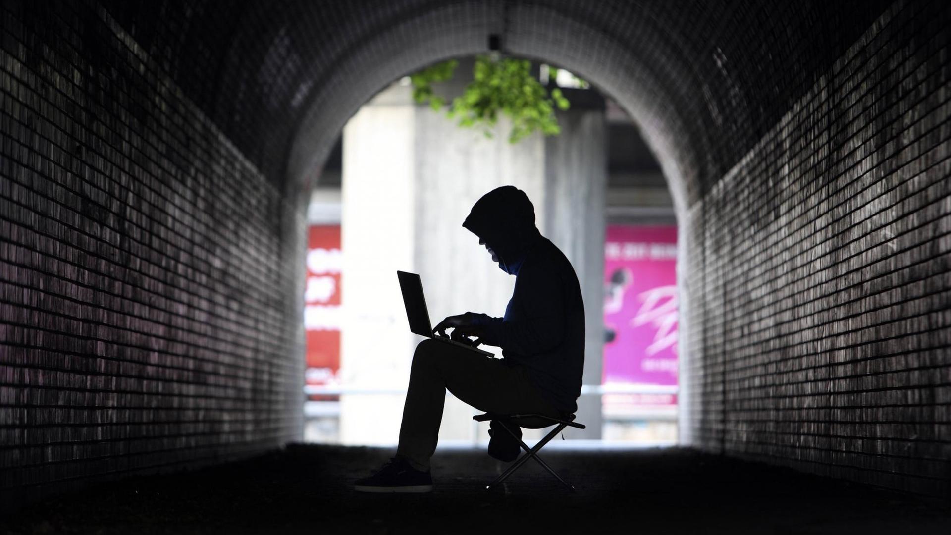 Mann mit Kapuzenpullover sitzt im Schatten eines Tunnels an einem Laptop Mann mit Kapuzenpullover sitzt im Schatten eines Tunnels an einem Laptop