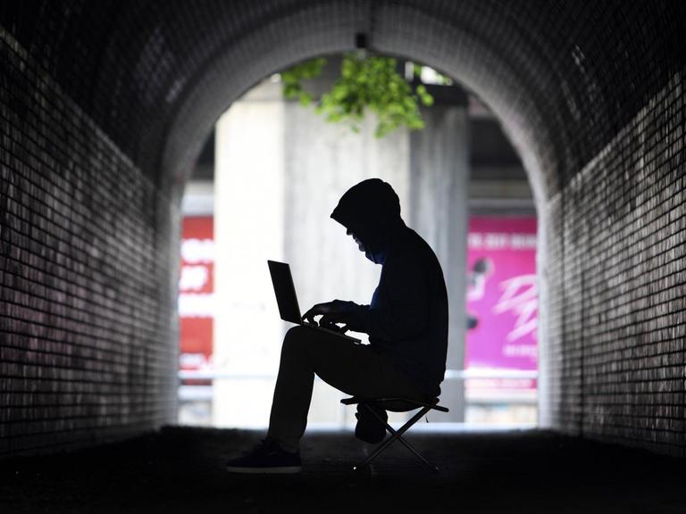 Mann mit Kapuzenpullover sitzt im Schatten eines Tunnels an einem Laptop