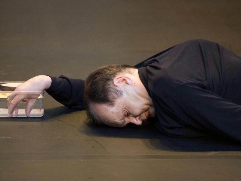 Der Tänzer Raimund Hoghe liegt auf dem Boden und taucht seine rechte Hand in eine durchsichtige Schale, die mit Wasser gefüllt ist.