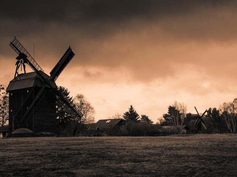 Zwei historische Windmühlen vor einem dramatisch gefärbten Himmel, im Vordergrund ist eine Wiese zu sehen.