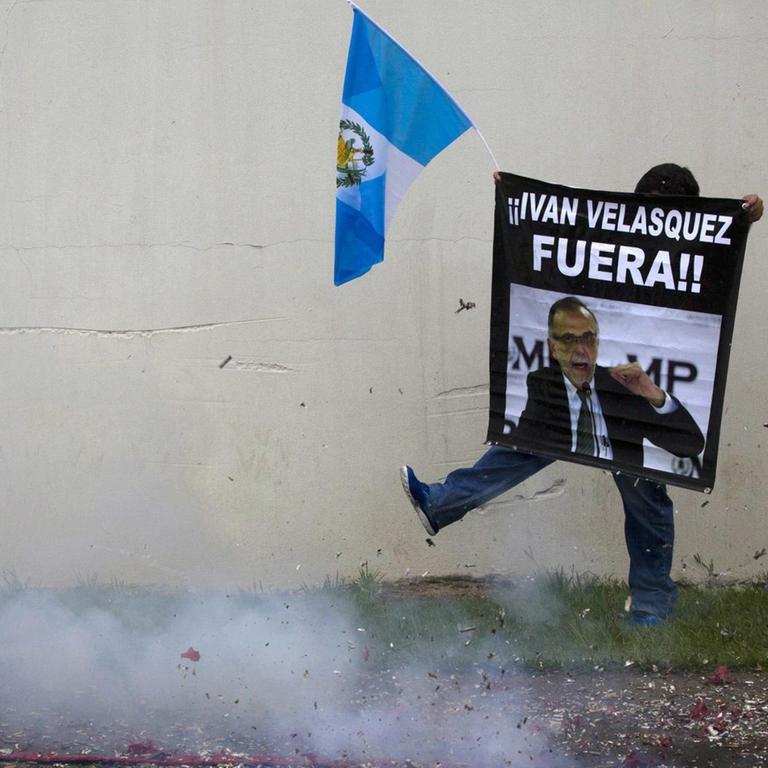 Ein Mann mit einem Protestplakat in Guatemala, vor ihm der Rauch eines Feuerwerkskörpers.