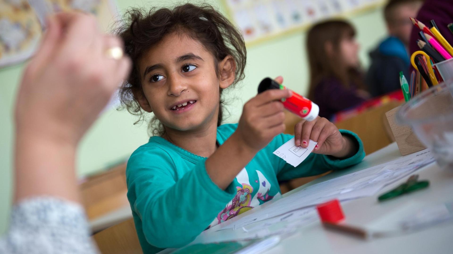 Ein Mädchen mit dunklen Locken arbeitet mit einem Klebestift und lächelt die Lehrerin an. Ein Mädchen mit dunklen Locken arbeitet mit einem Klebestift und lächelt die Lehrerin an.