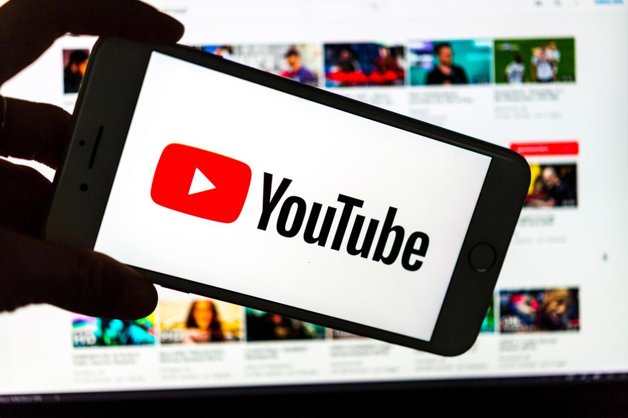 Das Logo des Video-Portals YouTube wird auf dem Display eines Smartphones angezeigt. Im Hintergrund ist auf einem Bildschirm die YouTube Homepage zu sehen. 