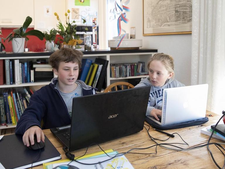 Zwei Jugendliche sitzen in einem Wohnzimmer vor Laptops und machen Hausaufgaben.