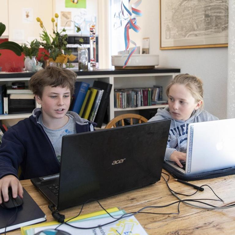Zwei Jugendliche sitzen in einem Wohnzimmer vor Laptops und machen Hausaufgaben.