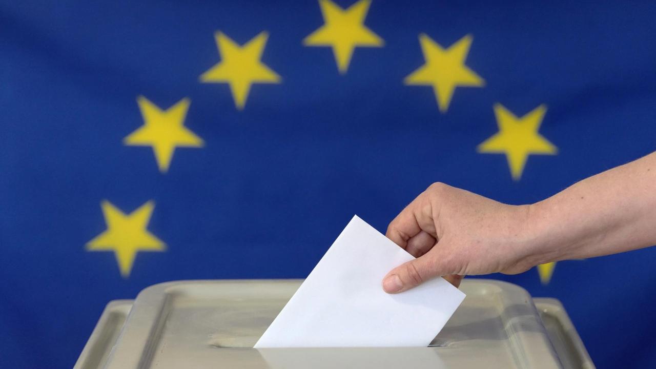 Eine Hand steckt einen Umschlag in eine Wahlurne vor der Europafahne. Symbolfoto. Eine Hand steckt einen Umschlag in eine Wahlurne vor der Europafahne. Symbolfoto.