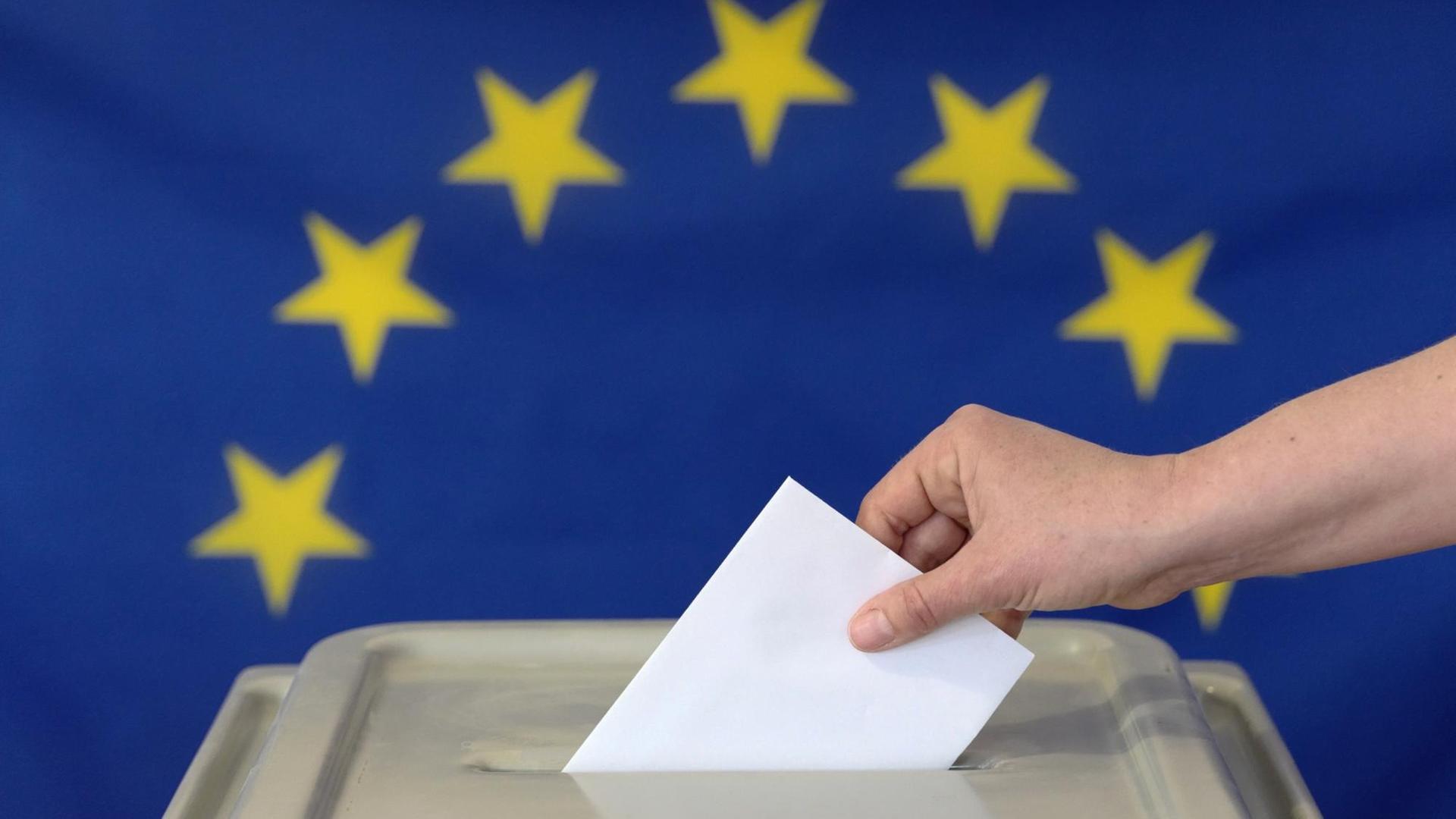 Eine Hand steckt einen Umschlag in eine Wahlurne vor der Europafahne. Symbolfoto. Eine Hand steckt einen Umschlag in eine Wahlurne vor der Europafahne. Symbolfoto.