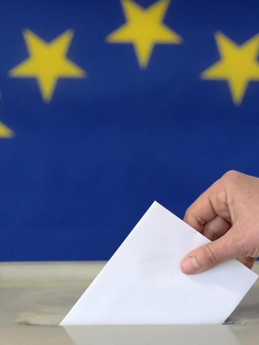 Eine Hand steckt einen Umschlag in eine Wahlurne vor der Europafahne. Symbolfoto.
