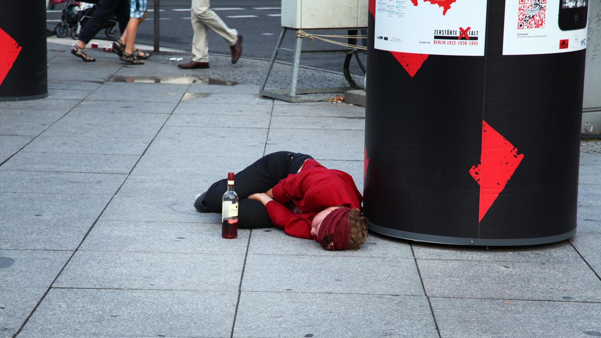Eine junge Frau liegt völlig betrunken auf dem Pflaster an einer Plakatsäule. Vor ihr steht steht eine halb ausgetrunkene Flasche Rotwein.