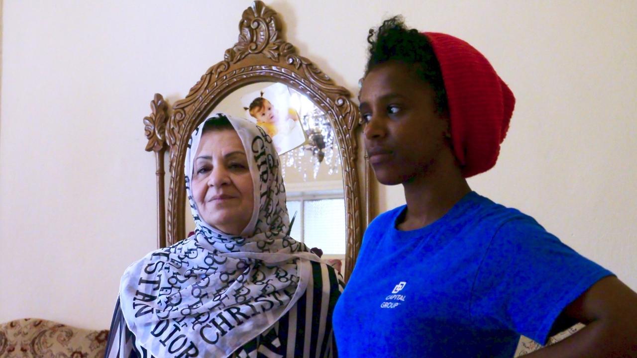 Eine junge Frau (namentlich Zola; im Bild rechts) mit T-Shirt und Mütze steht neben einen älteren Frau, ihrer Dienstherrin (links), in Gewand und Kopftuch Eine junge Frau (namentlich Zola; im Bild rechts) mit T-Shirt und Mütze steht neben einen älteren Frau, ihrer Dienstherrin (links), in Gewand und Kopftuch
