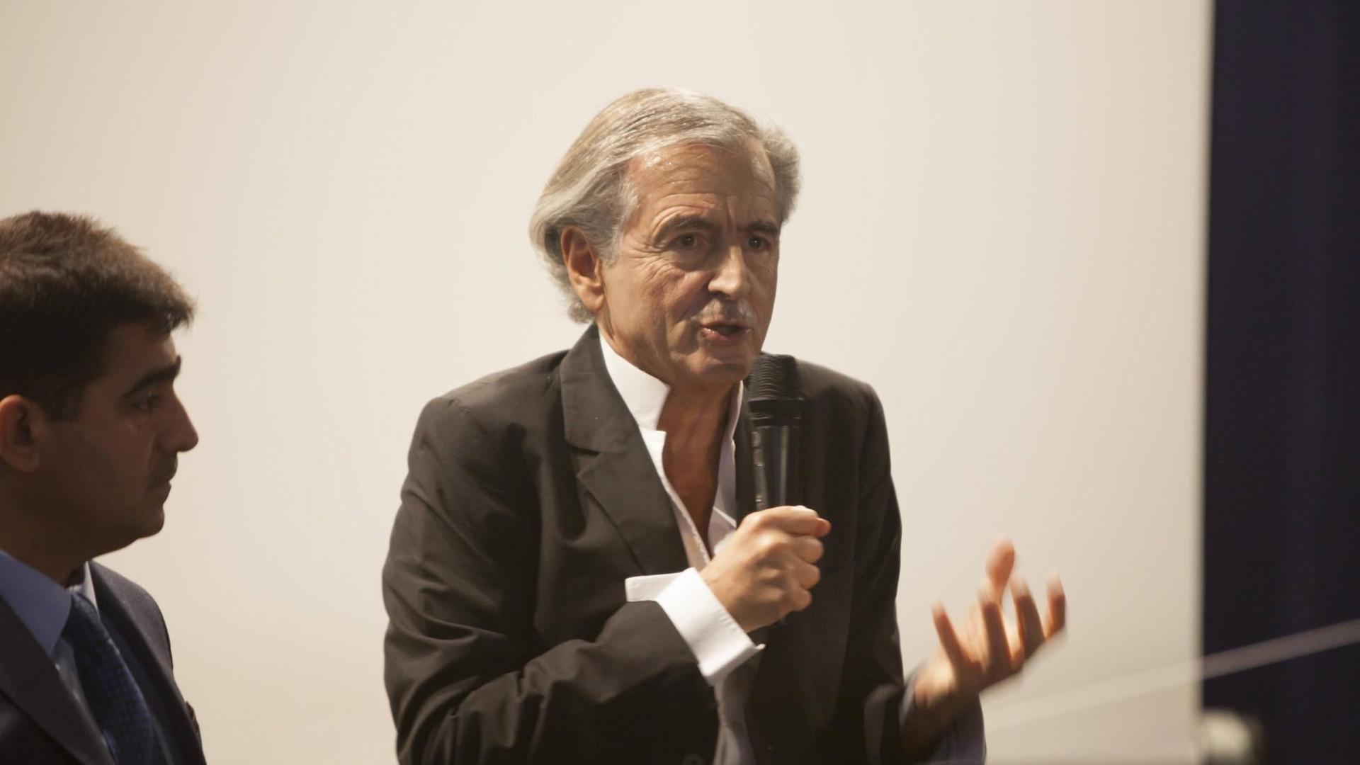 der Philosoph Bernard-Herni Levy steht auf einem Podium und spricht in ein Mikrofon, er gestikuliert mit seiner linken Hand. der Philosoph Bernard-Herni Levy steht auf einem Podium und spricht in ein Mikrofon, er gestikuliert mit seiner linken Hand.
