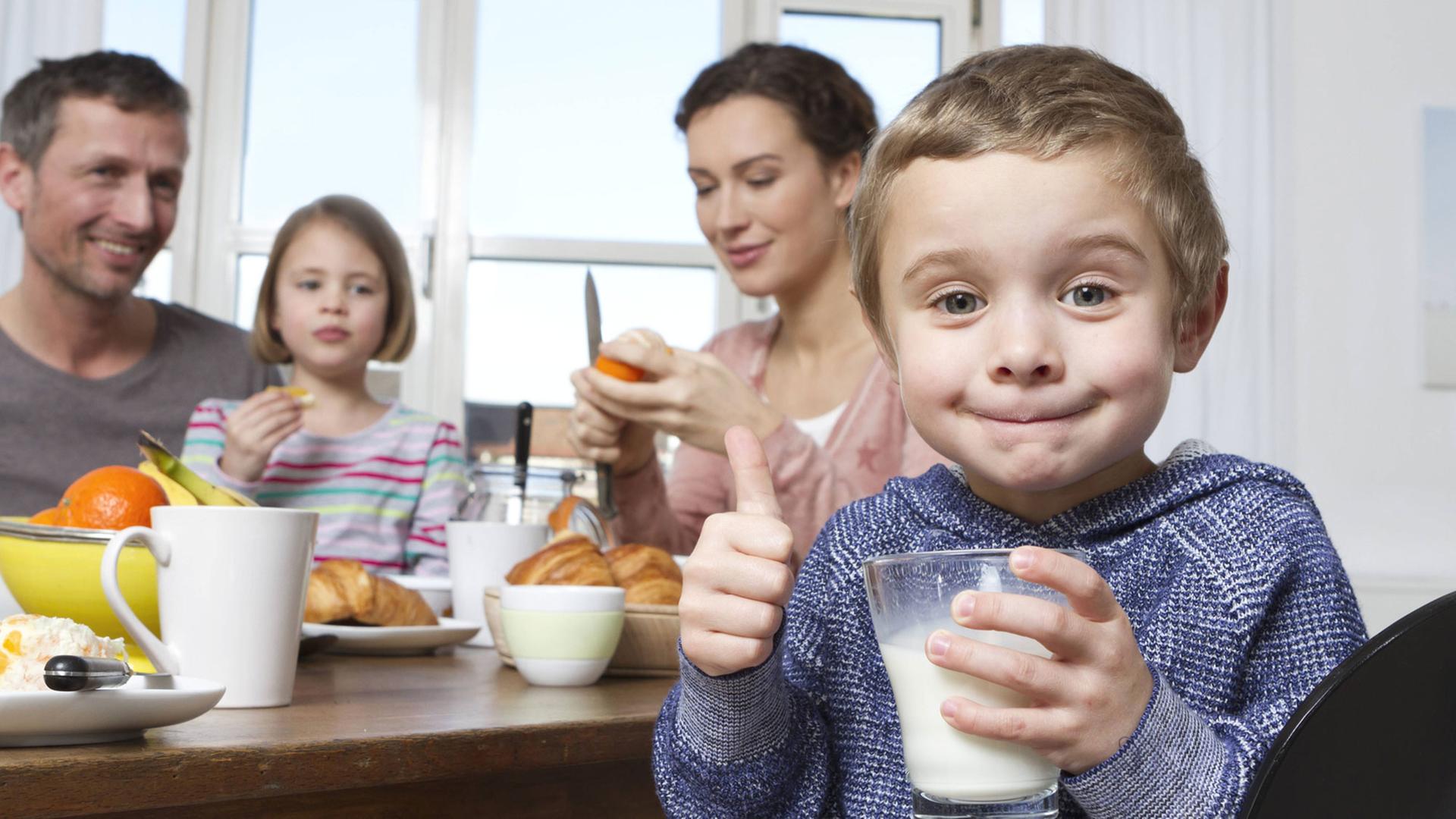 Mutter, Vater, Tochter und Sohn beim Frühstück. Der Sohn hält ein Glas Milch in der Hand und reckt den Daumen in die Kamera. Mutter, Vater, Tochter und Sohn beim Frühstück. Der Sohn hält ein Glas Milch in der Hand und reckt den Daumen in die Kamera.