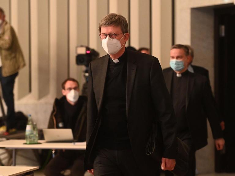 Kardinal Woelki kommt zur Presse-Konferenz. Im Hinter-Grund sind Kameras.