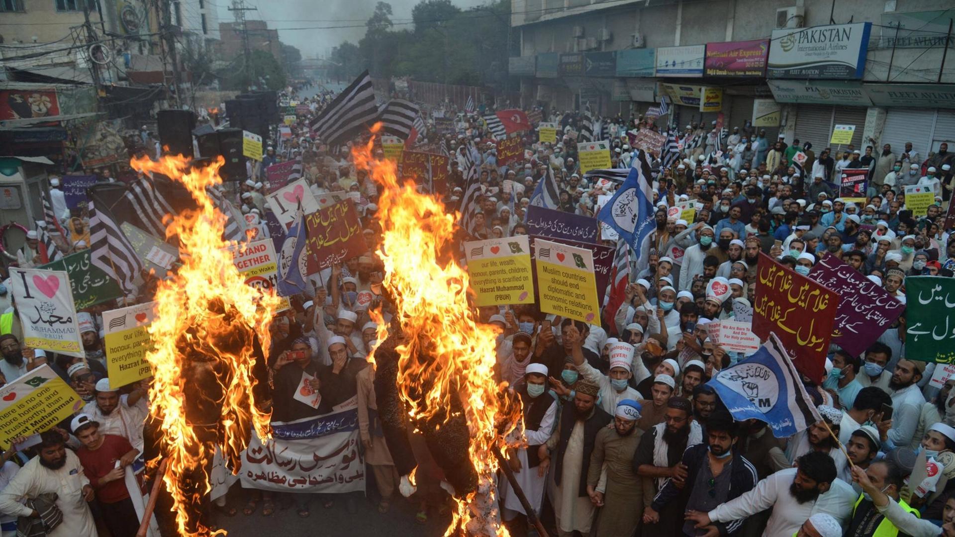 Dicht gedrängt stehen Menschen in Pakistan bei einer Straßendemonstration. In der ersten Reihe schlagen Flammen hoch, weil die Protestierenden Plakate verbrennen. Dicht gedrängt stehen Menschen in Pakistan bei einer Straßendemonstration. In der ersten Reihe schlagen Flammen hoch, weil die Protestierenden Plakate verbrennen.