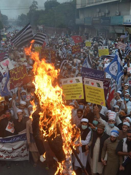 Dicht gedrängt stehen Menschen in Pakistan bei einer Straßendemonstration. In der ersten Reihe schlagen Flammen hoch, weil die Protestierenden Plakate verbrennen.