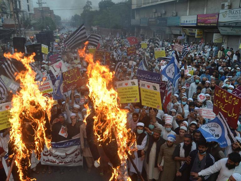 Dicht gedrängt stehen Menschen in Pakistan bei einer Straßendemonstration. In der ersten Reihe schlagen Flammen hoch, weil die Protestierenden Plakate verbrennen.