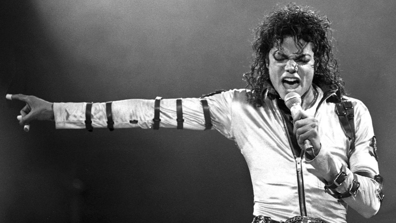 Schwarzweißaufnahme von Michael Jackson bei einem Live-Auftritt in den 80ern: Der weißgekleidete Sänger steht frontal vor der Kamera, singt in ein Mikrofon und weist mit dem rechten Arm streng zur Seite.