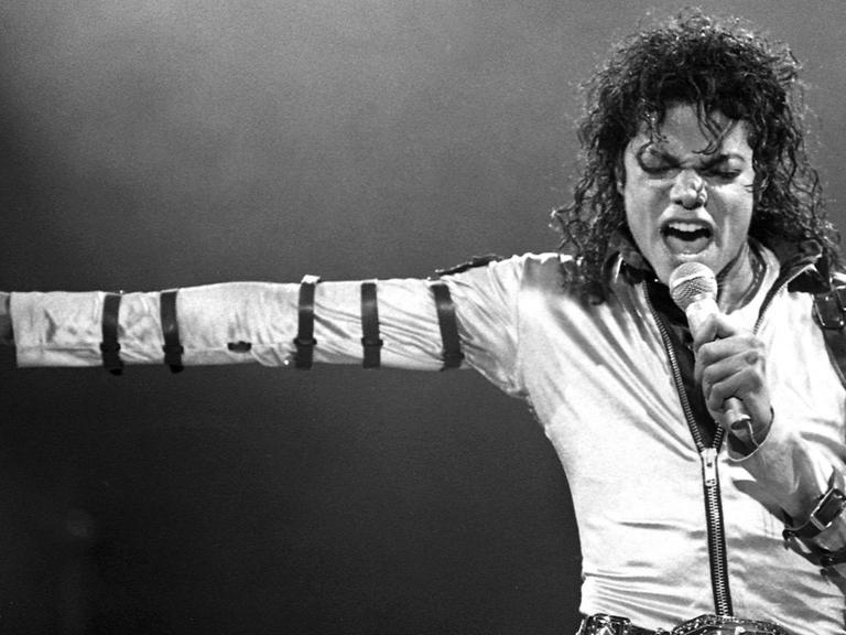 Schwarzweißaufnahme von Michael Jackson bei einem Live-Auftritt in den 80ern: Der weißgekleidete Sänger steht frontal vor der Kamera, singt in ein Mikrofon und weist mit dem rechten Arm streng zur Seite.